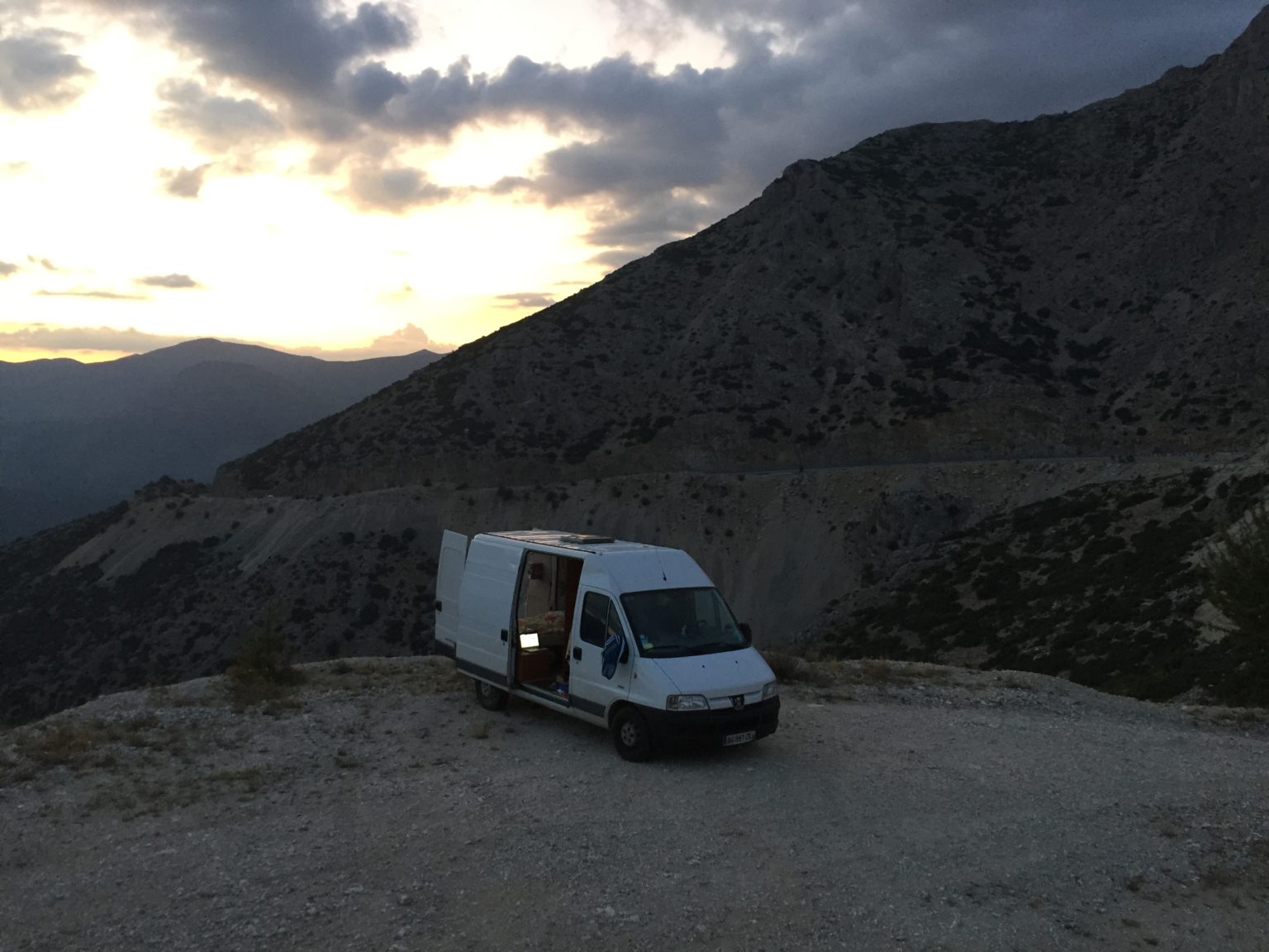 Campervan parked on a mountain overlook at sunset, with dramatic clouds and rugged terrain in the background.