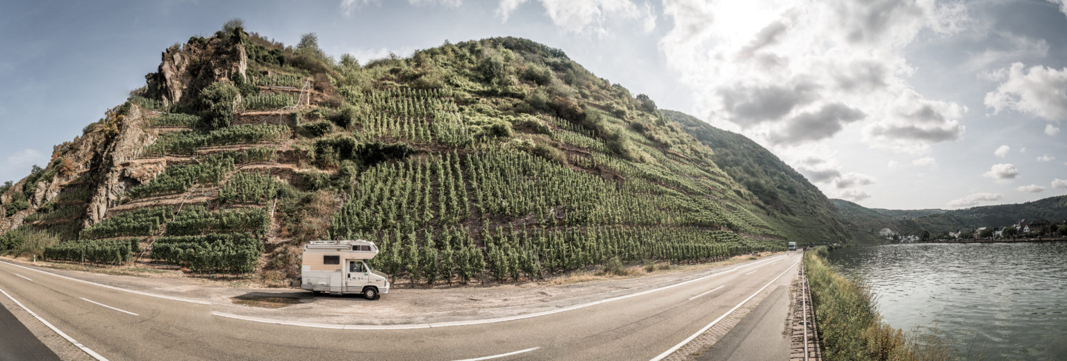 Campervan parked by a scenic vineyard hillside, near a river under a partly cloudy sky.