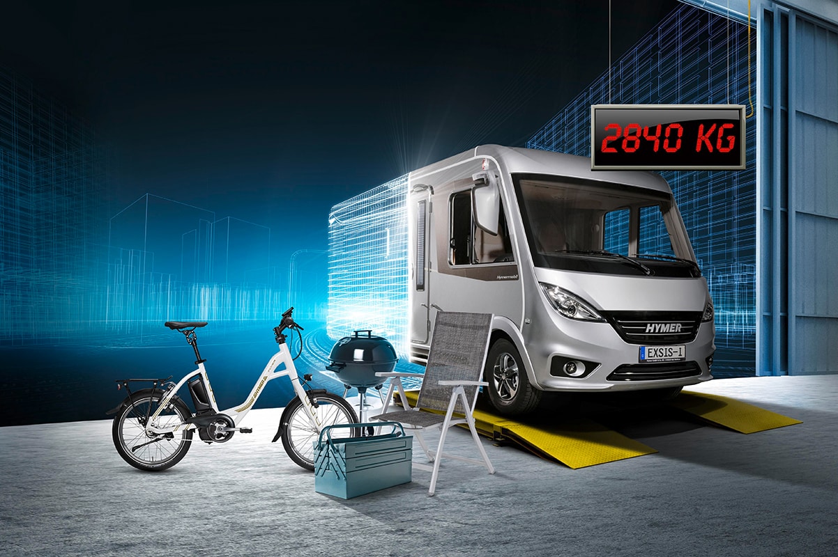 Camping-car Hymer sur balance, entouré de vélo, barbecue et chaises, ambiance moderne et technologique.