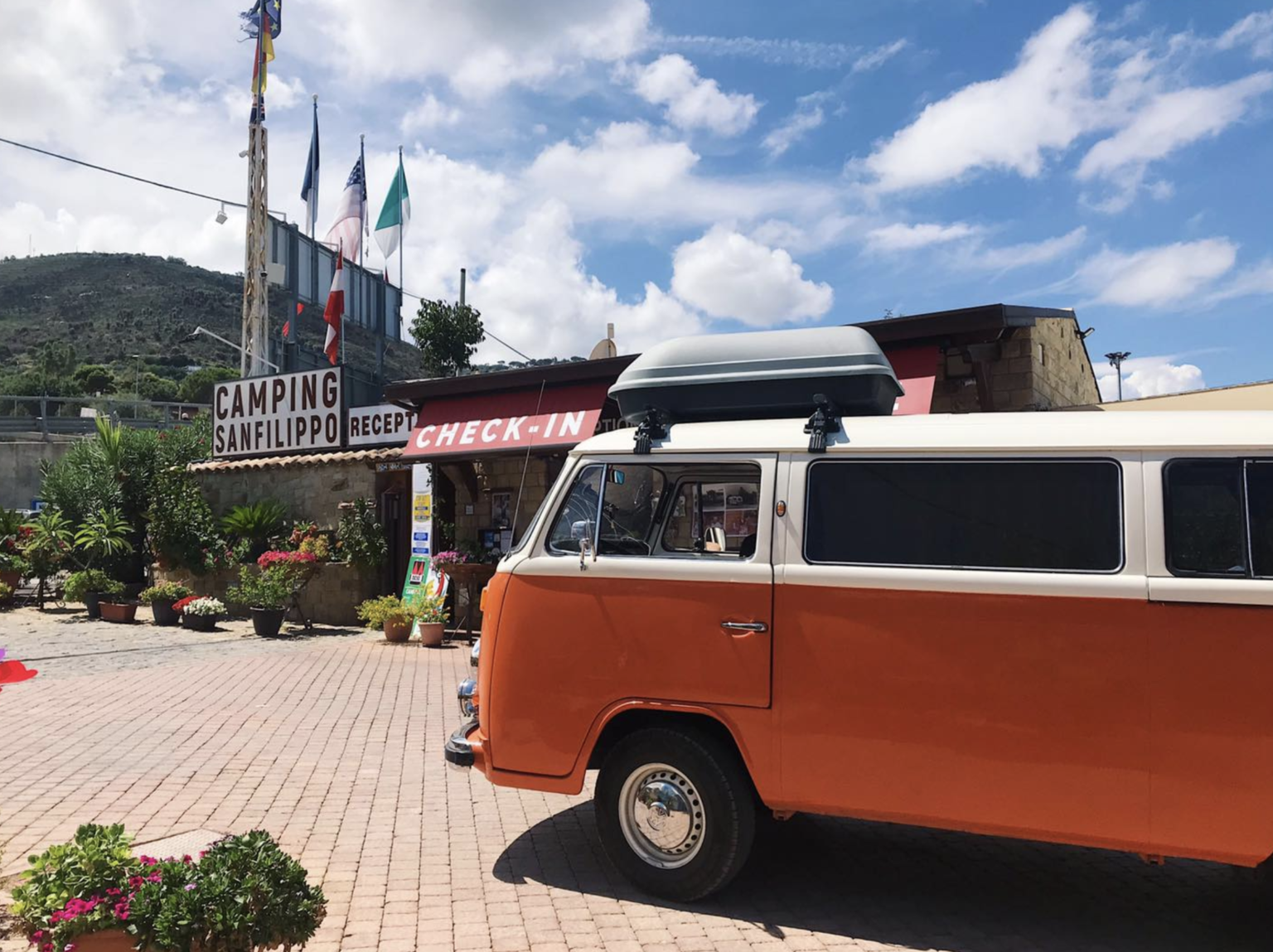 Orange campervan parked at Camping Sanfilippo entrance under a blue sky, with flags and lush greenery around.