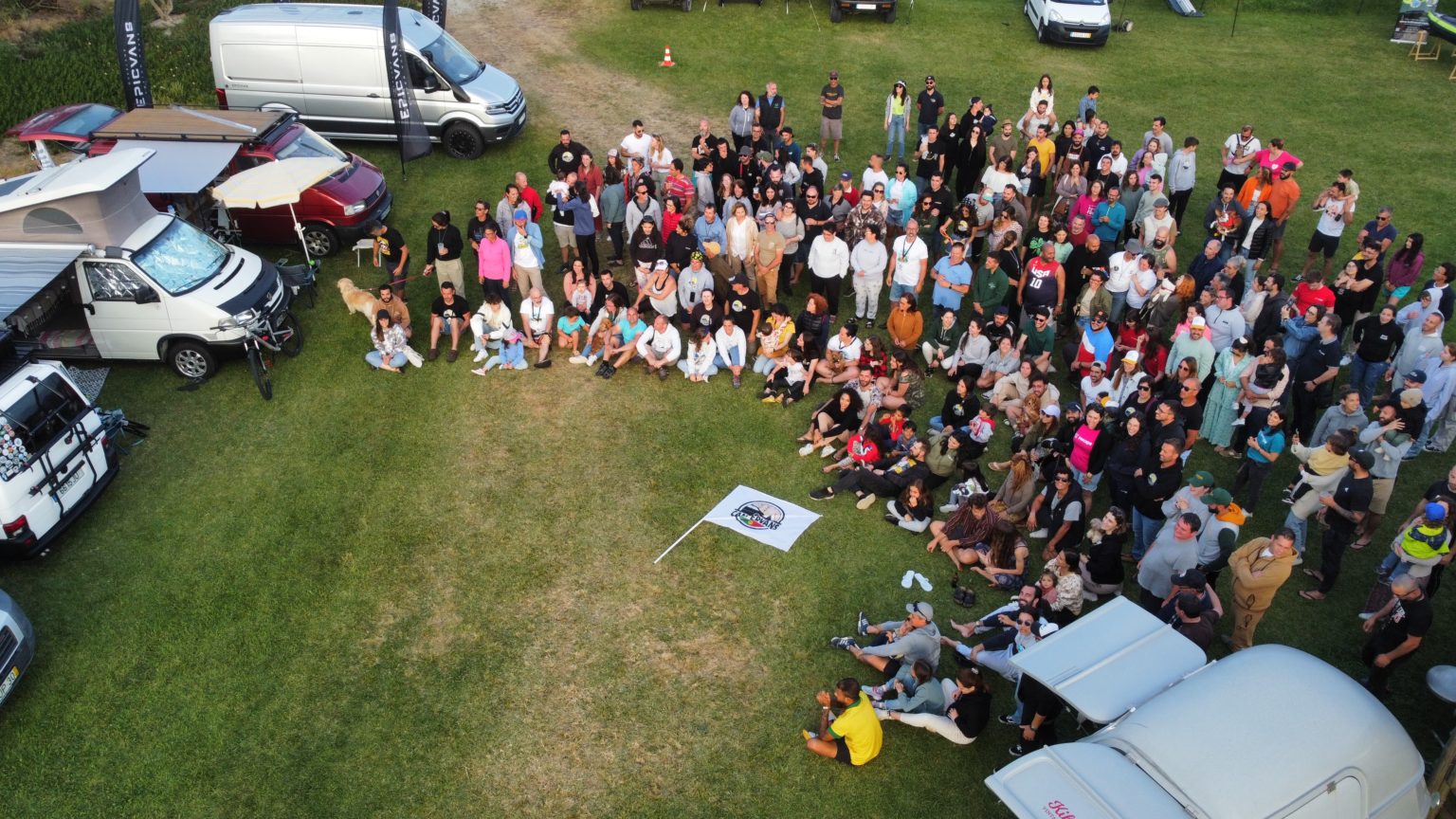 Large group of people gathered on a grassy field with campervans, creating a lively and communal atmosphere.