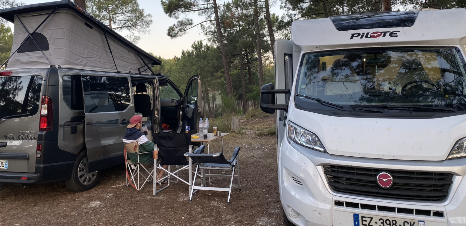 Camping en forêt avec un van et un camping-car, ambiance détendue autour de chaises pliantes et d'une table.