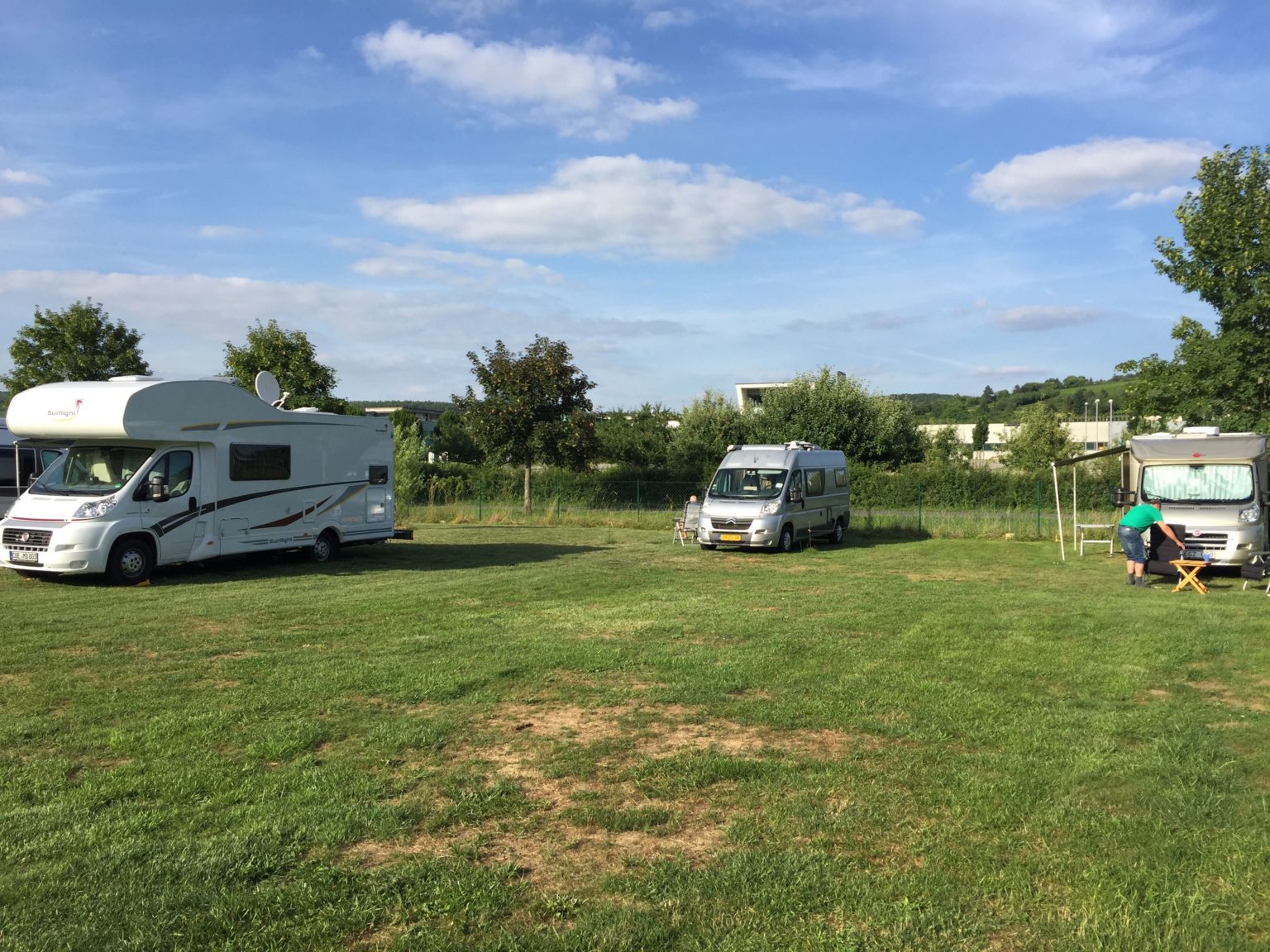 Motorhomes parked on a grassy field under a blue sky; a person sits nearby enjoying the view.
