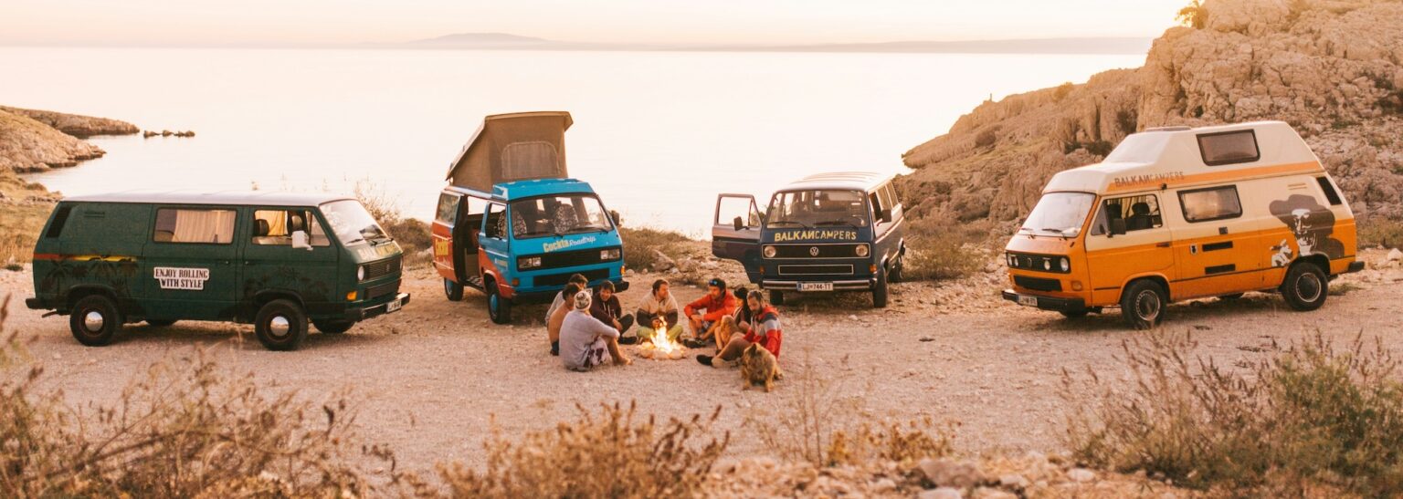 Campers gather around a beach bonfire, surrounded by colorful vans at sunset, creating a warm, adventurous vibe.