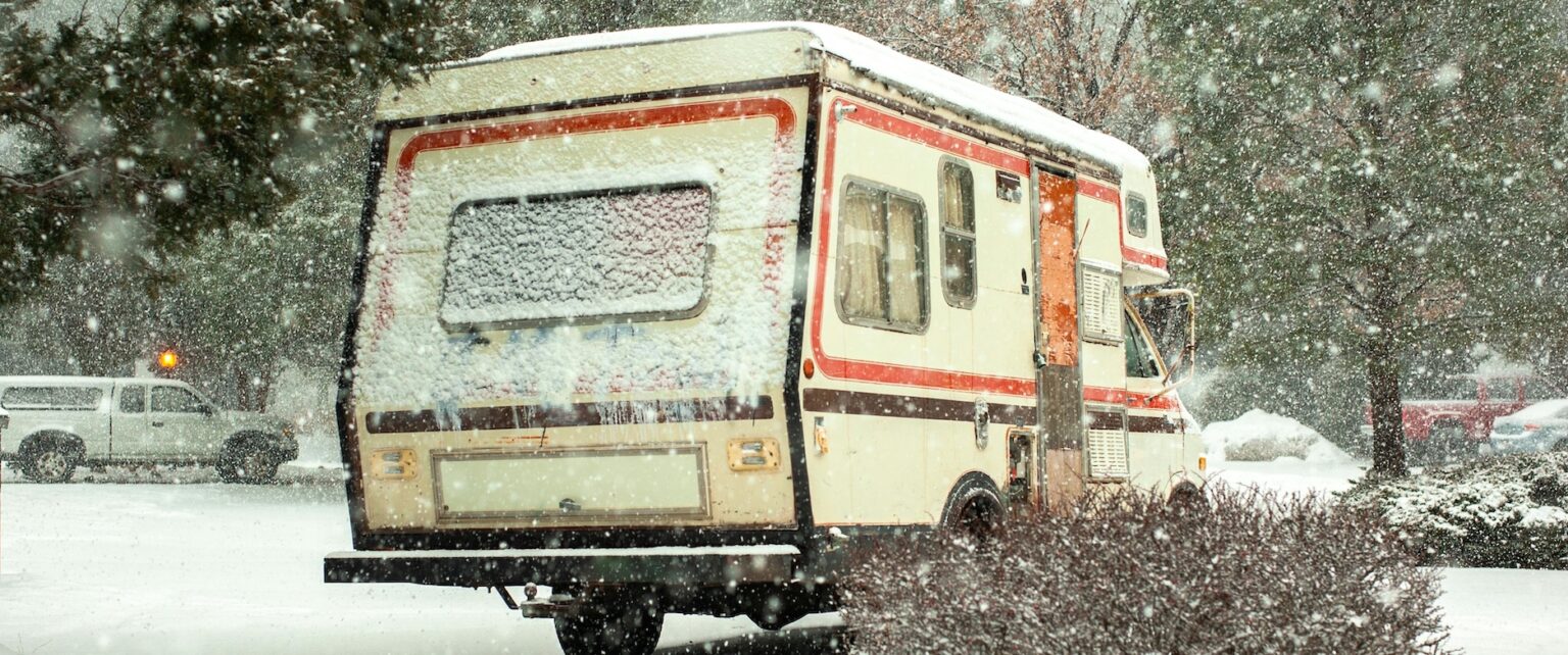 Camper innevato parcheggiato in un paesaggio invernale, circondato da alberi e neve cadente.