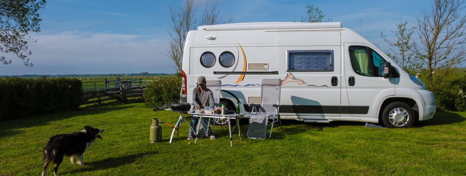 Camper parcheggiato in un prato verde, con sedie da campeggio e un cane che gioca all'aperto. Atmosfera rilassata.