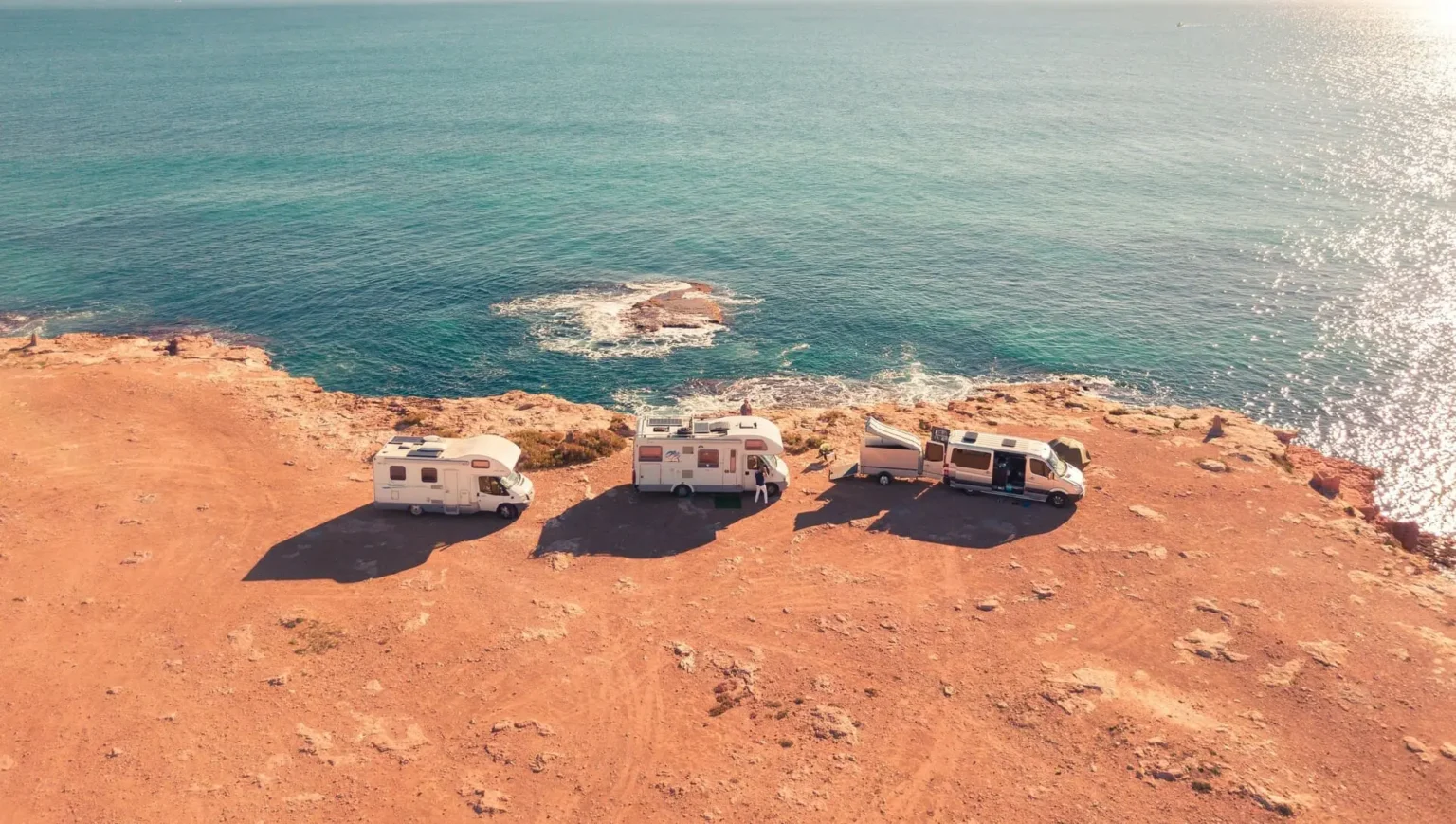 Three campervans parked on a rocky coastal cliff overlooking the ocean under a clear blue sky.