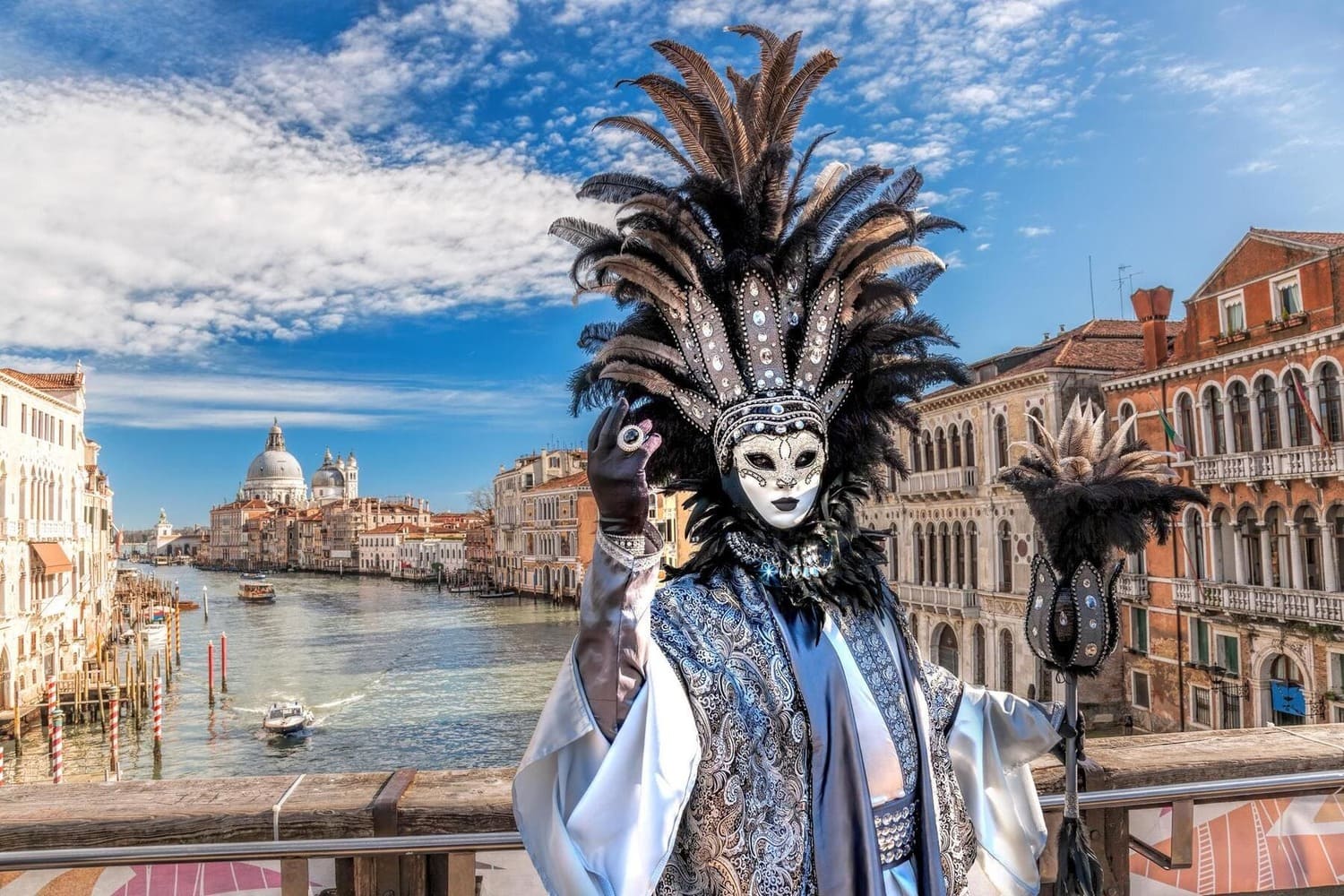 Personne en costume vénitien sur un pont à Venise, avec le Grand Canal en arrière-plan sous un ciel bleu.