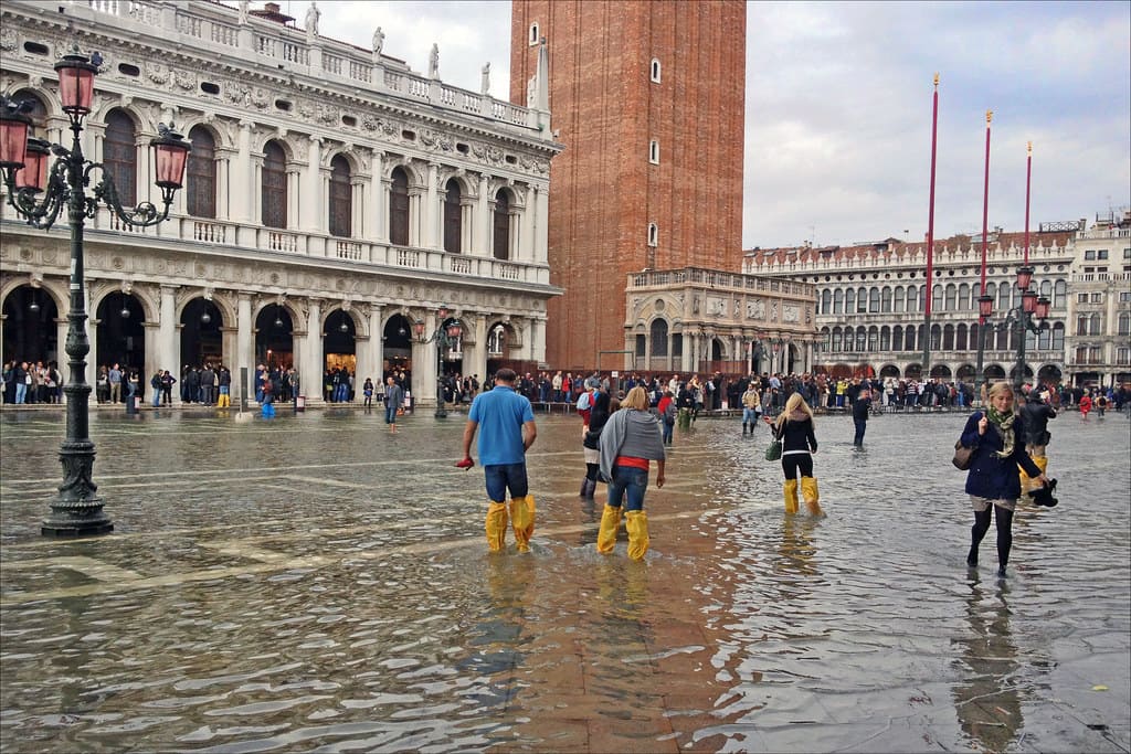 Personnes marchant avec des bottes dans une place inondée à Venise, ambiance animée et touristique.