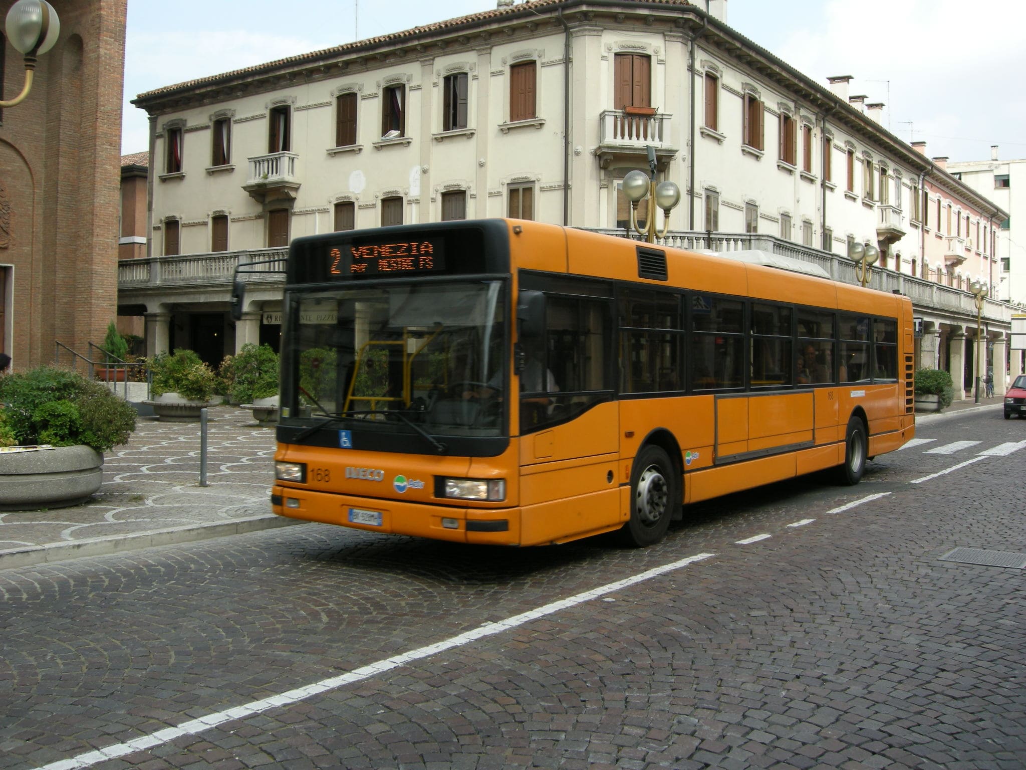 Bus orange en route vers Venise, traversant une rue pavée bordée de bâtiments historiques. Ambiance urbaine et touristique.