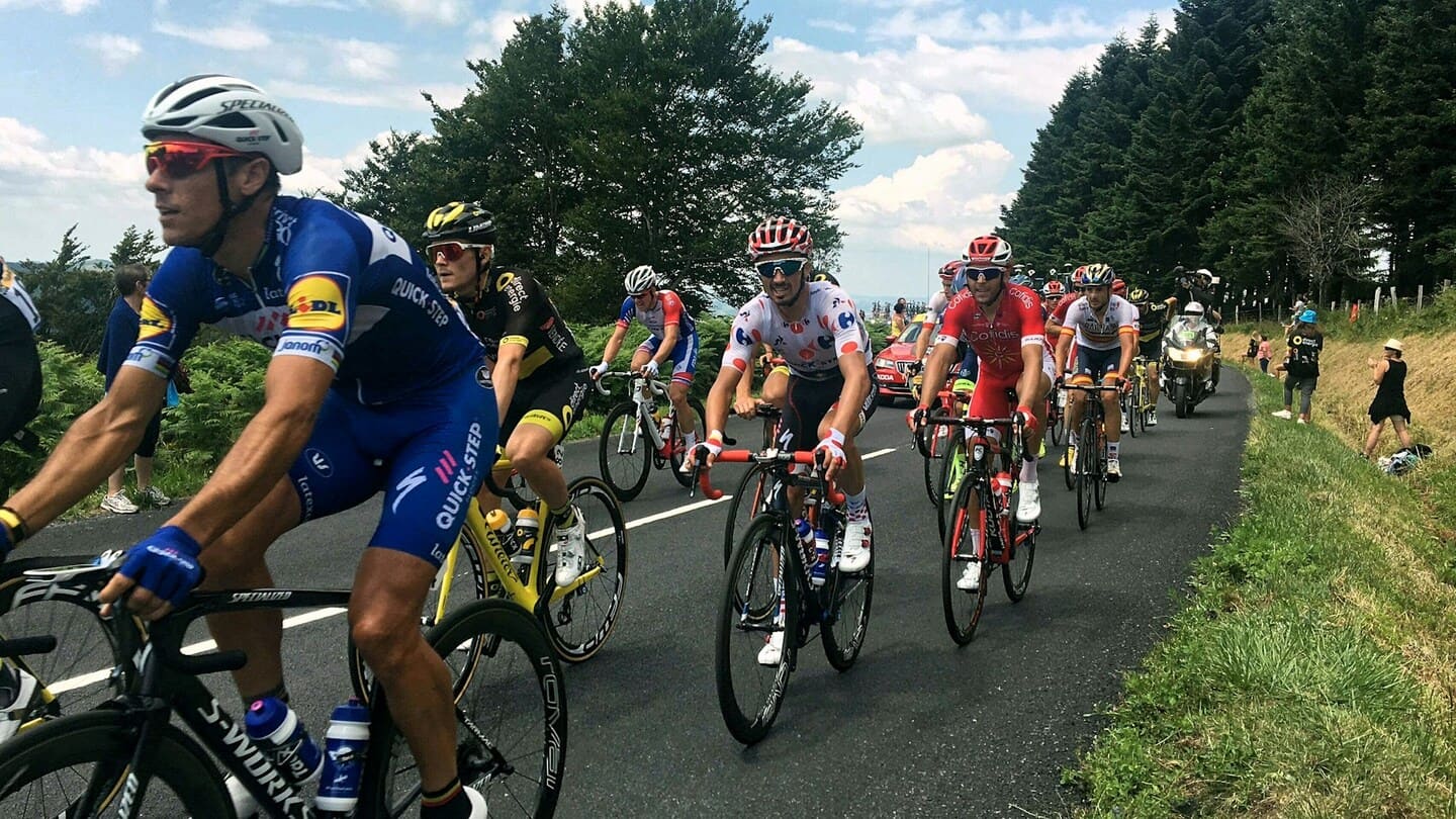 Peloton du Tour de France roulant sur une route de campagne entourée de forêt et de spectateurs.